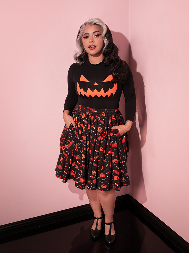 Full length shot of female model wearing the TRICK R TREAT™ Skater Skirt in Candy Corn Novelty Print with black long sleeved pumpkin shirt and black flats.