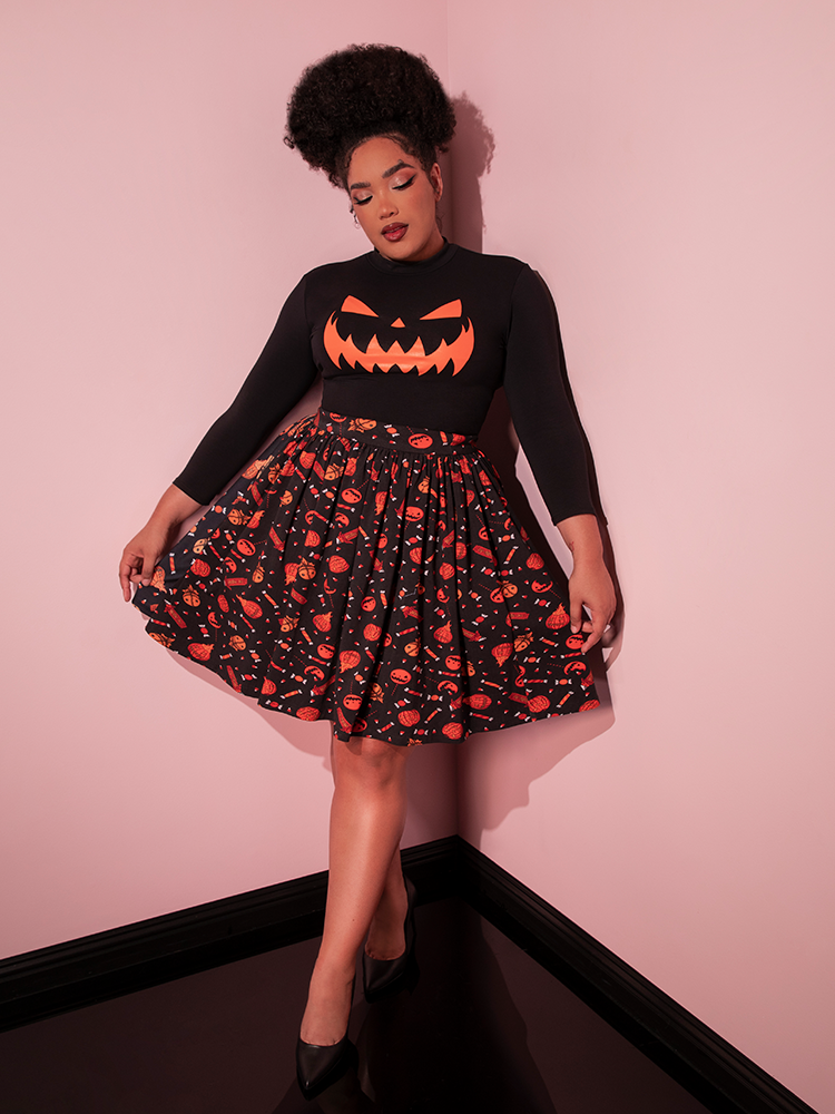 Female model pulls out the sides of the TRICK R TREAT™ Skater Skirt in Candy Corn Novelty Print to display the colorful pumpkin and candy print design.