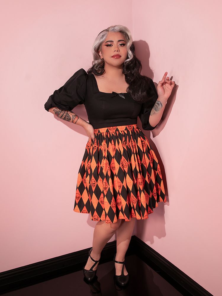 Female model with silver and black hair posing with her hand up in the air and the other held on her hip wearing the TRICK R TREAT™ Skater Skirt in Halloween Harlequin Print.