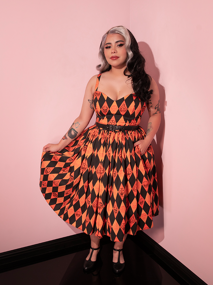 Model poses with on hand tucked into a deep side pocket of her TRICK R TREAT™ Sweetheart Swing Dress in Halloween Harlequin Print and the other holding out the side of the skirt to show off the black and orange print.