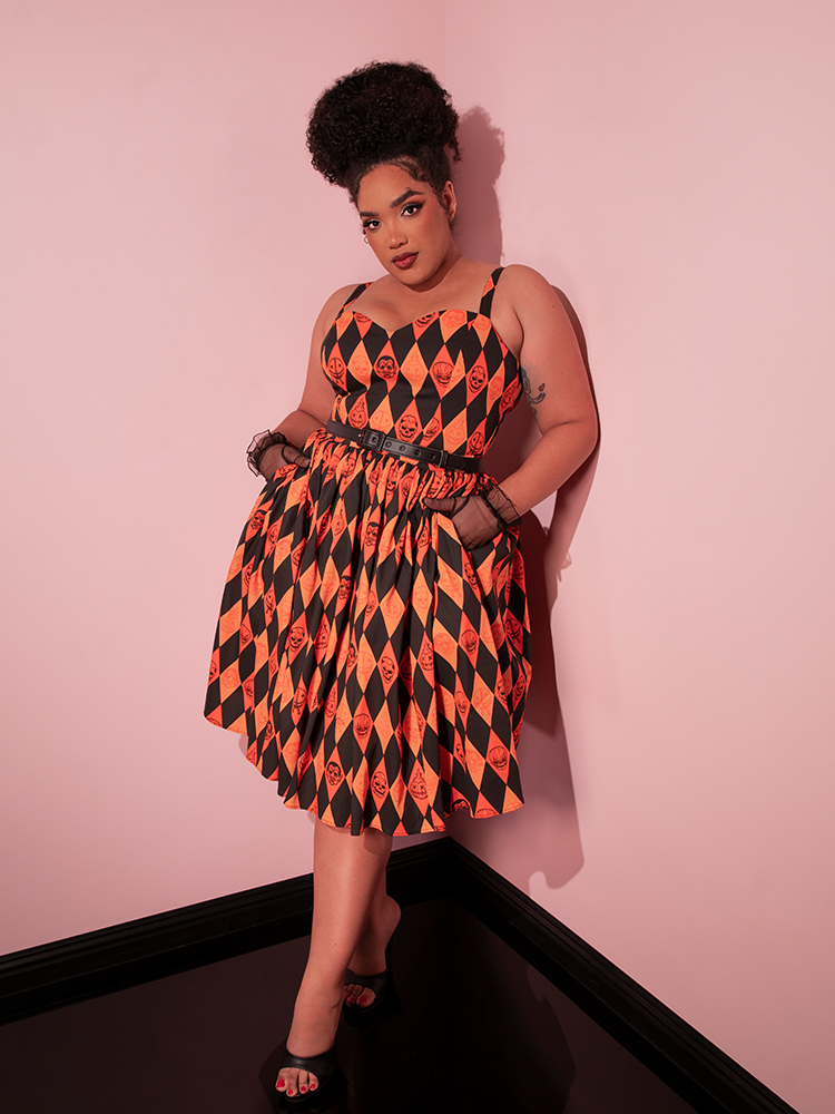 Model tucking her hands into the pockets of the TRICK R TREAT™ Sweetheart Swing Dress in Halloween Harlequin Print while leaning back against a wall. 