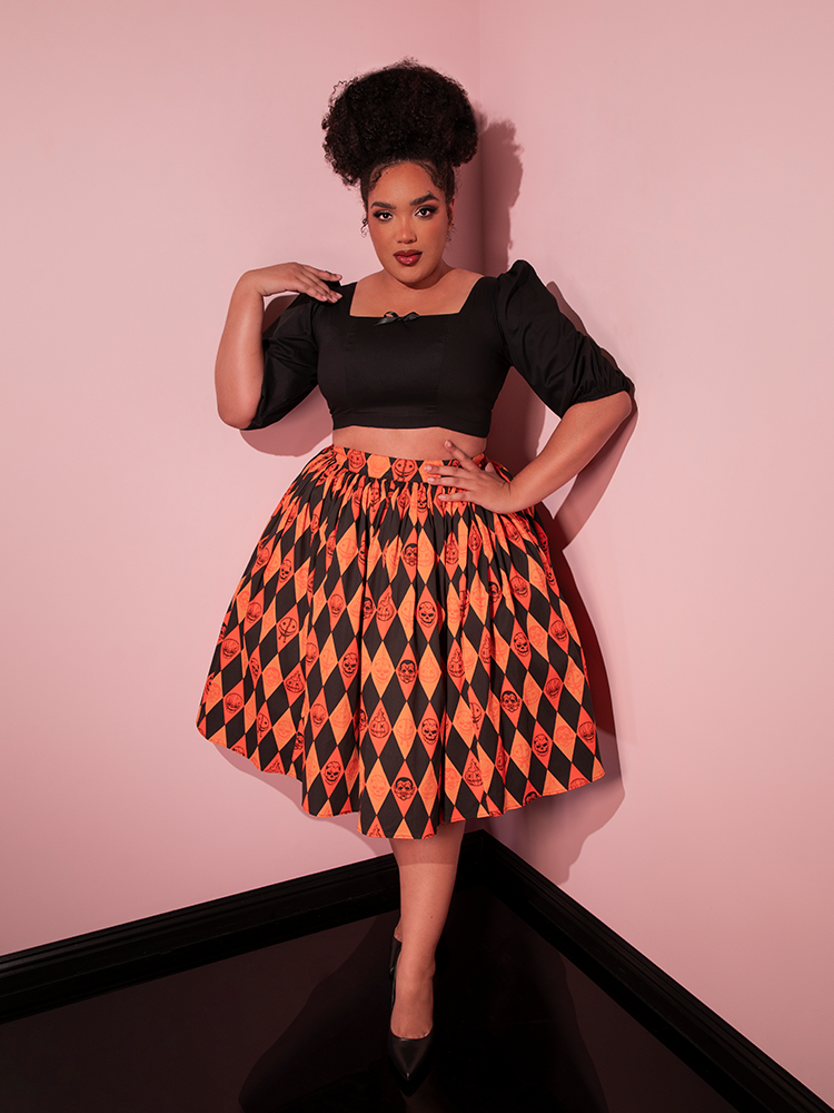 Full length shot of model standing in the corner of a room wearing a retro outfit including the TRICK R TREAT™ Swing Skirt in Halloween Harlequin Print and black top.