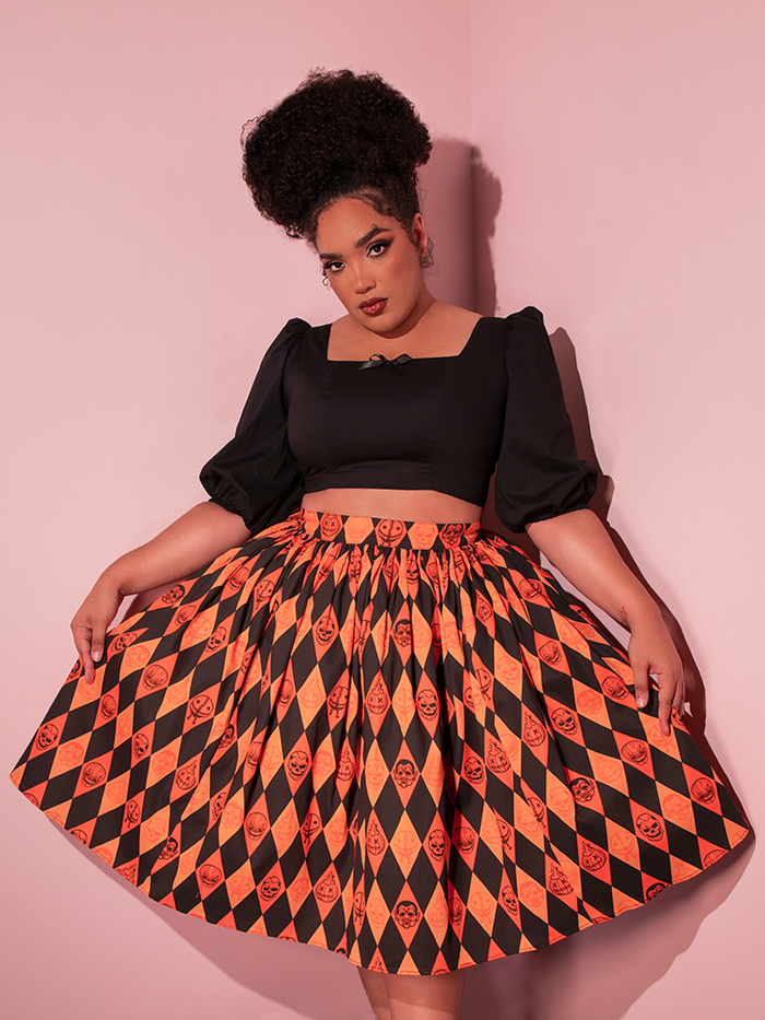Model posing in a black retro top and holding out the sides of her TRICK R TREAT™ Swing Skirt in Halloween Harlequin Print.
