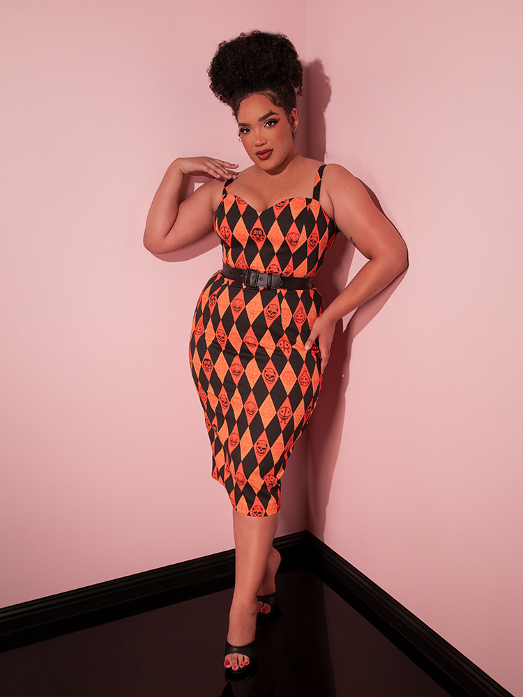 Model leaning against a pink wall while modeling the all new classic Halloween look vintage dress from Vixen Clothing - TRICK R TREAT™ Sweetheart Wiggle Dress in Halloween Harlequin Print.