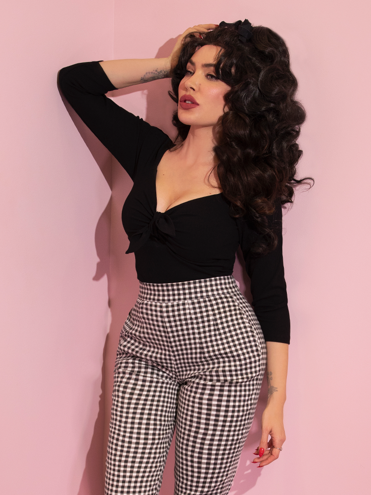 A closeup of Micheline Pitt looking away from camera modeling the tie me up top in black from Vixen Clothing paired with gingham pants.