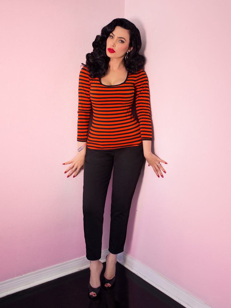 A full length shot of Micheline Pitt modeling the Troublemaker top in orange and black stripes untucked by Vixen Clothing.