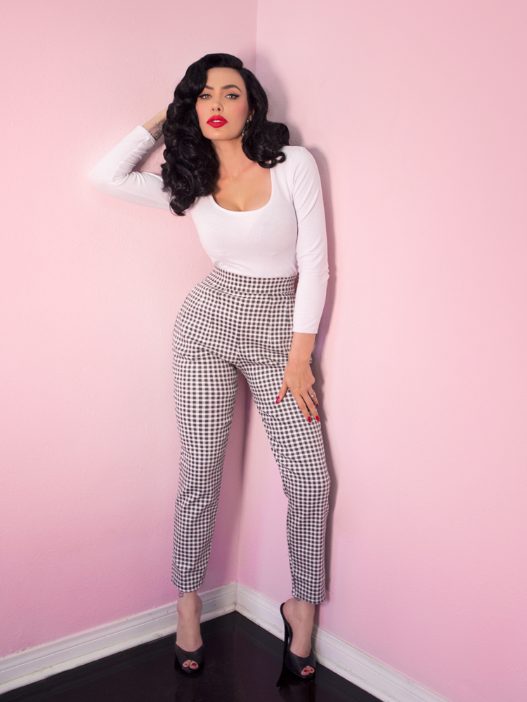 A full length shot of Micheline Pitt with her hand in her hair modeling the Troublemaker top in white paired with gingham cigarette pants.