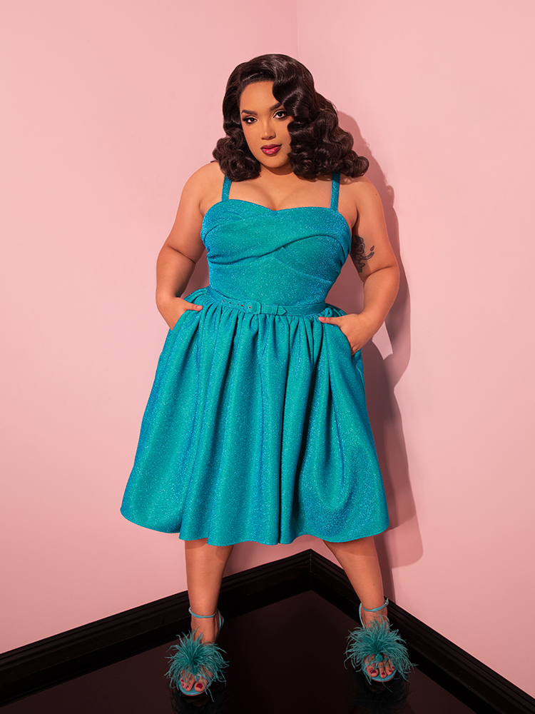 Commanding the spotlight, the female model strikes a captivating pose, highlighting the irresistible appeal of the Jawbreaker Swing Dress in Turquoise Lurex from Vixen Clothing, a celebrated brand specializing in retro dresses.