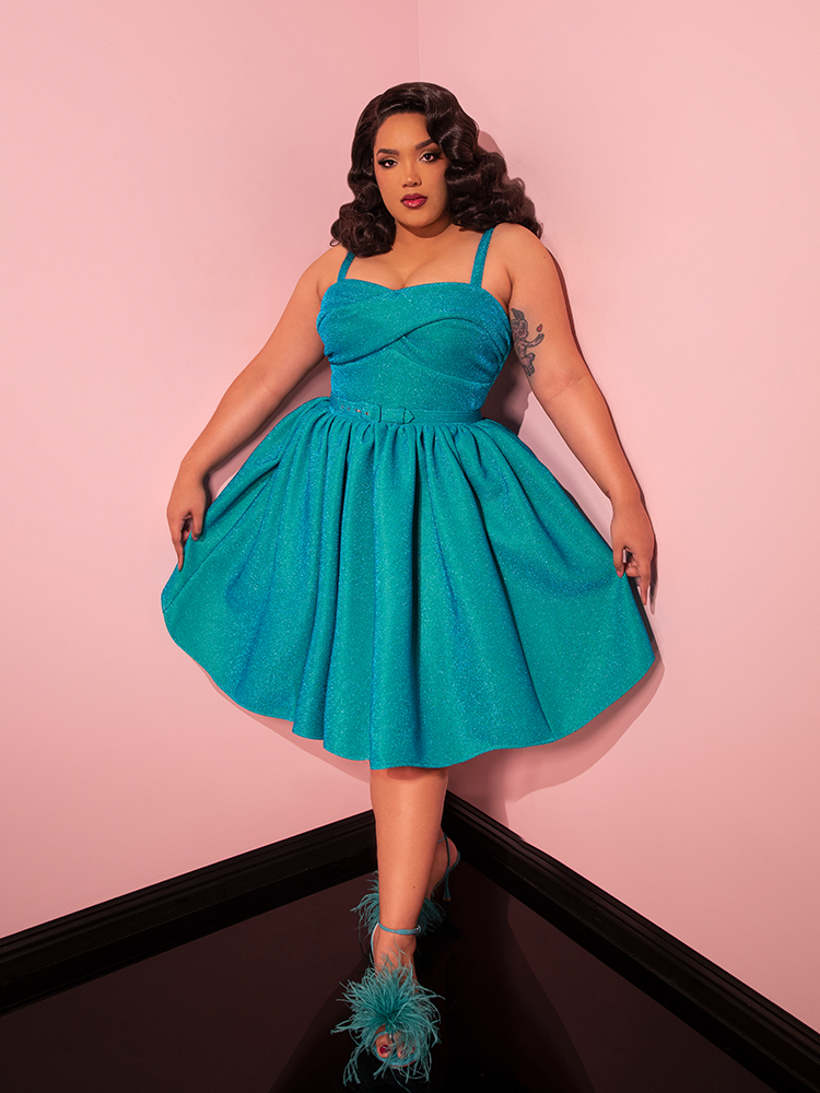 With undeniable allure, the female model confidently showcases the Turquoise Lurex Jawbreaker Swing Dress by Vixen Clothing, an enchanting garment that pays homage to the timeless charm of retro dresses.