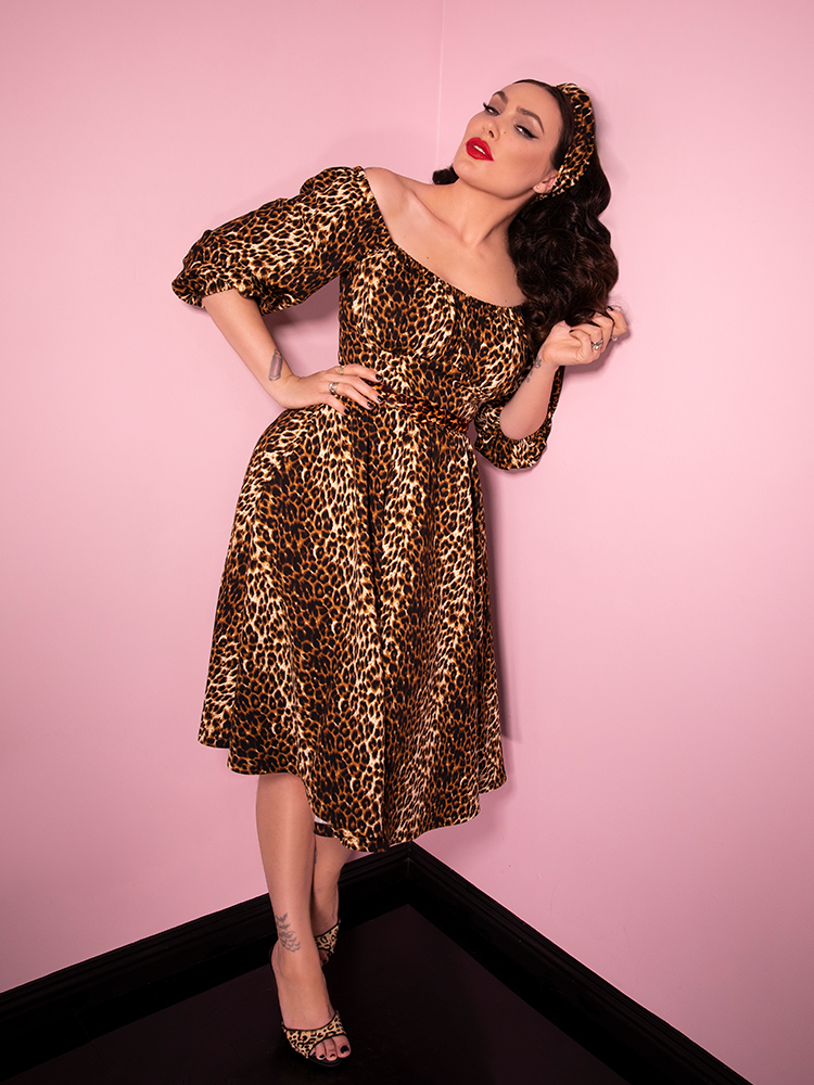 Leaning against a pink wall, Micheline Pitt models the Vacation Dress in Leopard Print from Vixen Clothing.
