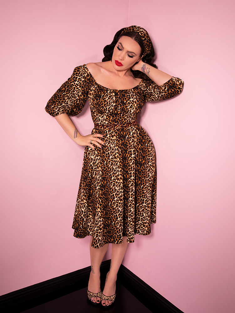 Micheline Pitt standing against pink painted walls and wearing the Vacation Dress in Vintage Leopard Print from Vixen Clothing. 