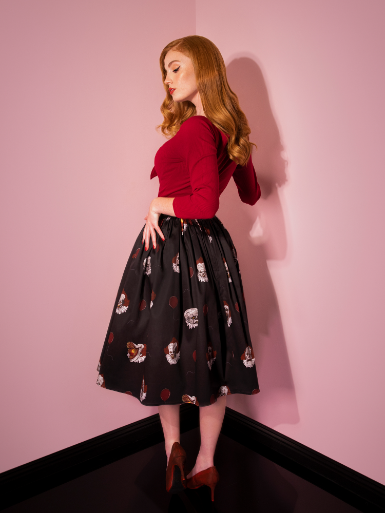 Turned away from the camera while looking down, Emily models the Pennywise swing skirt in black by Vixen Clothing.