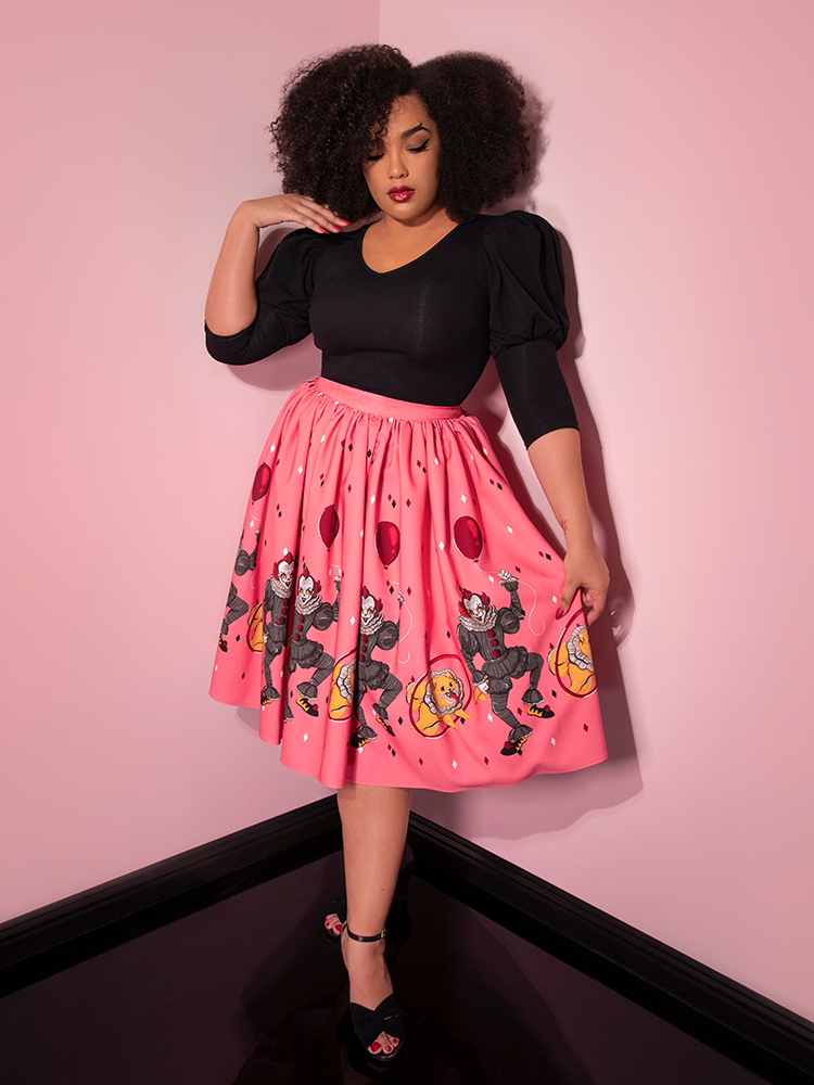 Looking down while holding her skirt, Ashleeta models the Dancing Clown swing skirt in pink by Vixen Clothing.