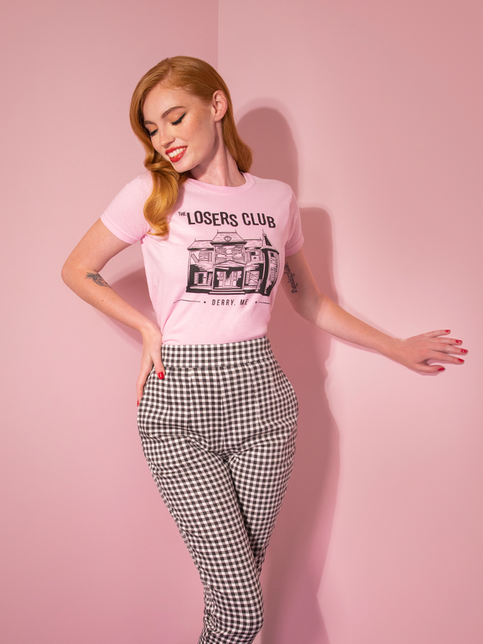 With her hand on her hip and looking down, model Emily wears the Losers Club pink tee from Vixen Clothing.