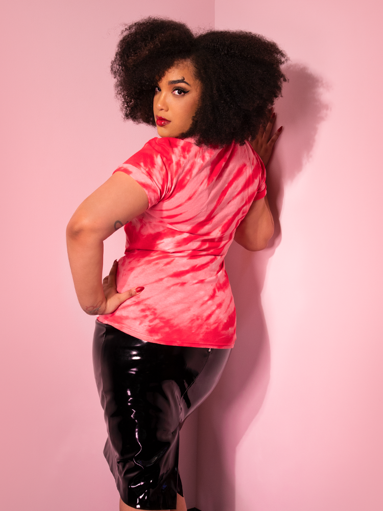 Turned away from the camera while leaning against a pink wall, Ashleeta models the Come Home red tie dye tee by Vixen Clothing.