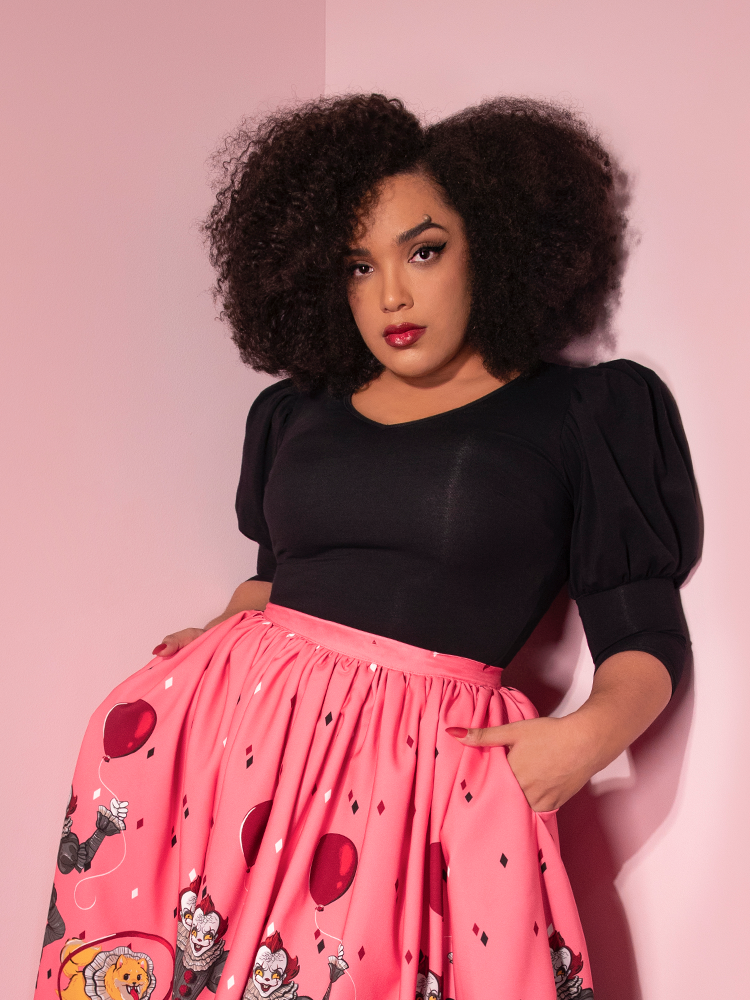 Looking into the camera with her hands in her pink skirt, Ashleeta models the Carnival top in black by Vixen Clothing.