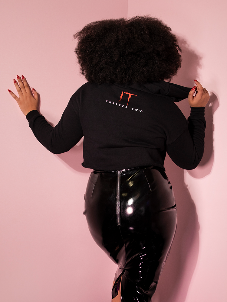 Turned away from the camera while leaning on a pink wall, Ashleeta models the back of the Home At Last cropped hoodie by Vixen Clothing.