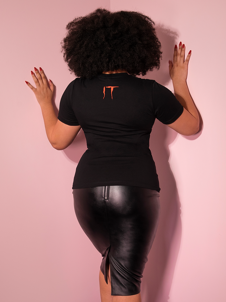 Turned away from the camera with her hands on the pink wall, Ashleeta models the back of the Pennywise float tee untucked by Vixen Clothing.