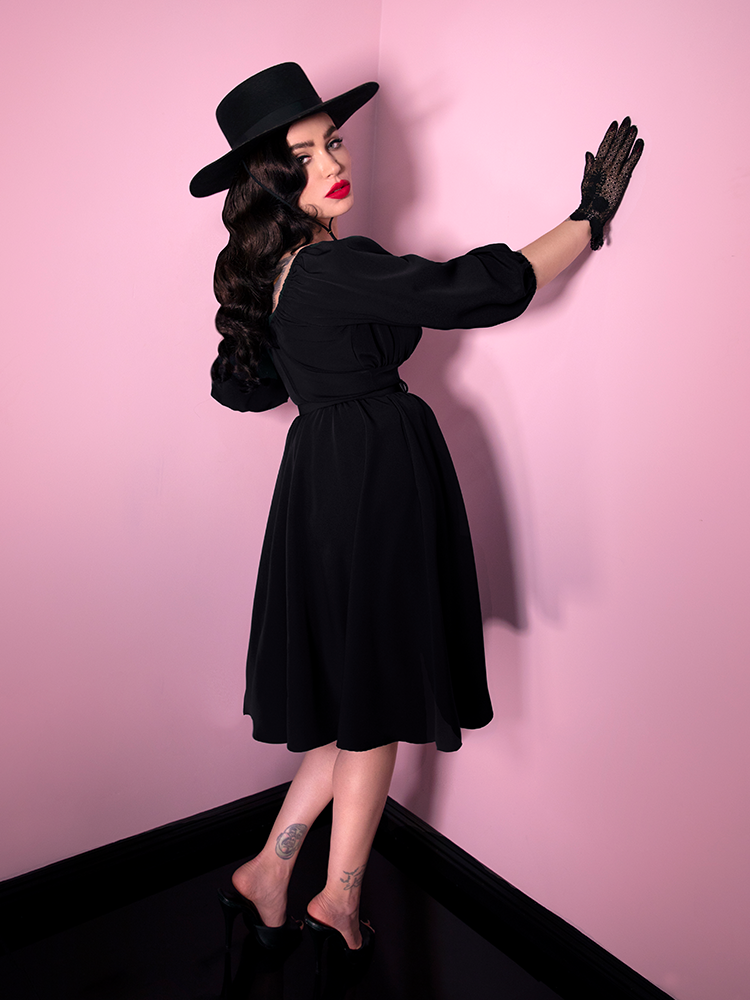 Facing a pink wall, Micheline Pitt is turning around to look off camera while wearing a retro style dress in black from Vixen Clothing.
