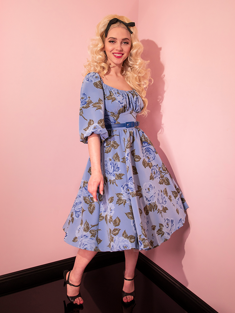 Sofia smiles while looking into the camera and wearing the Vacation Dress in Sunset Blue Roses from retro clothing brand Vixen Clothing.