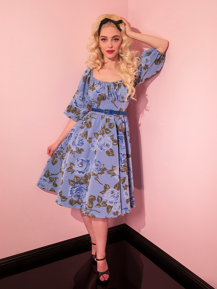 Sofia poses with one arm resting against the wall while wearing the Vacation Dress in Sunset Blue Roses. 