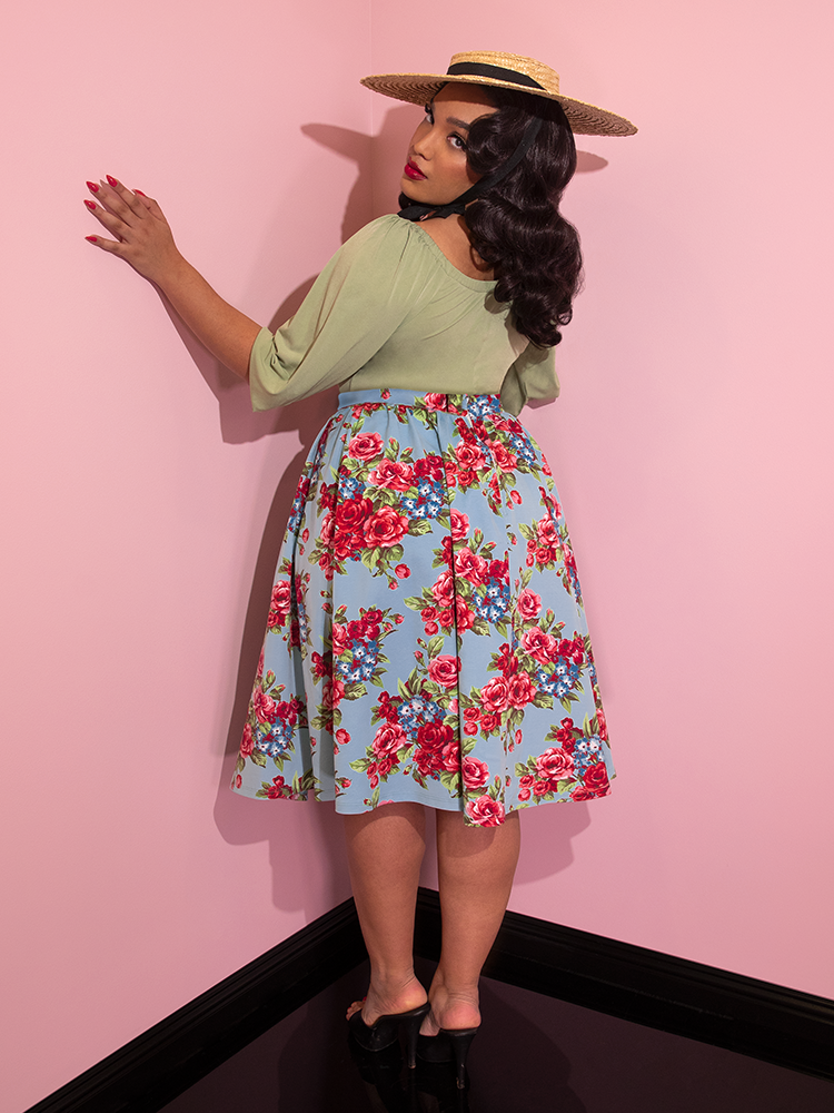 Ashleeta wearing a straw hat and green top looking over her shoulder modeling the Vacation skirt in blue and red rose print.
