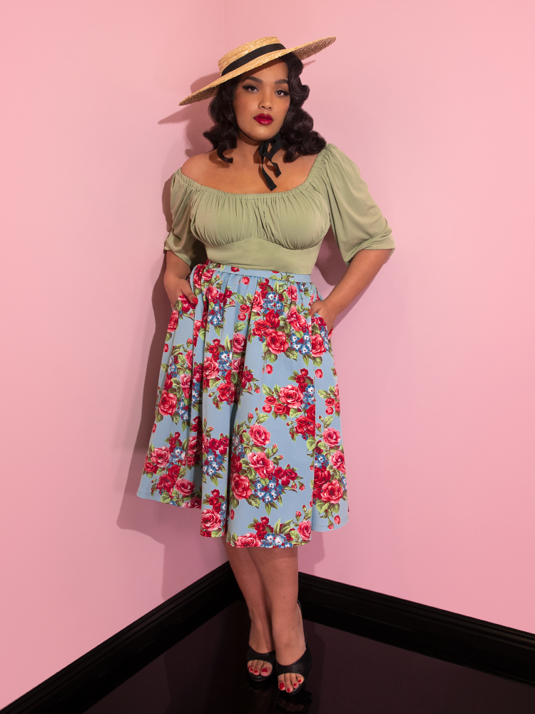 Ashleeta wearing a straw hat and green top modeling the Vacation skirt in blue and red rose print with her hands in her pockets.