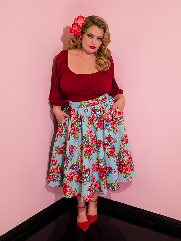 Blondie wearing flowers in her hair and a red top modeling the Vacation skirt in blue and red rose print with her hands in her pockets.