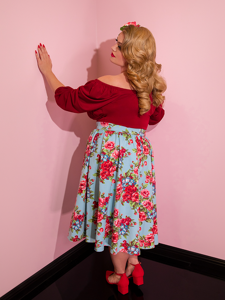 Blondie looking towards the wall wearing a red top modeling the Vacation skirt in blue and red rose print.