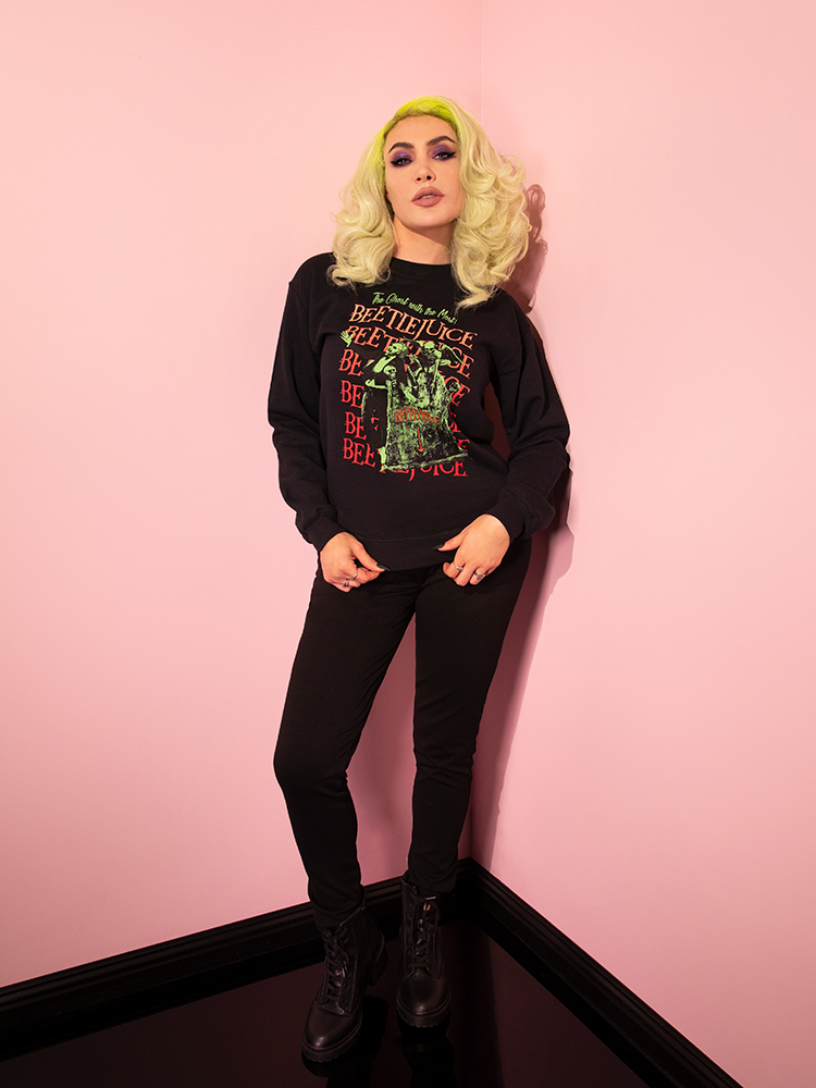 Micheline Pitt posing in the corner of a pink room wearing the Tombstone Sweatshirt with black jeans and black ankle length combat boots.