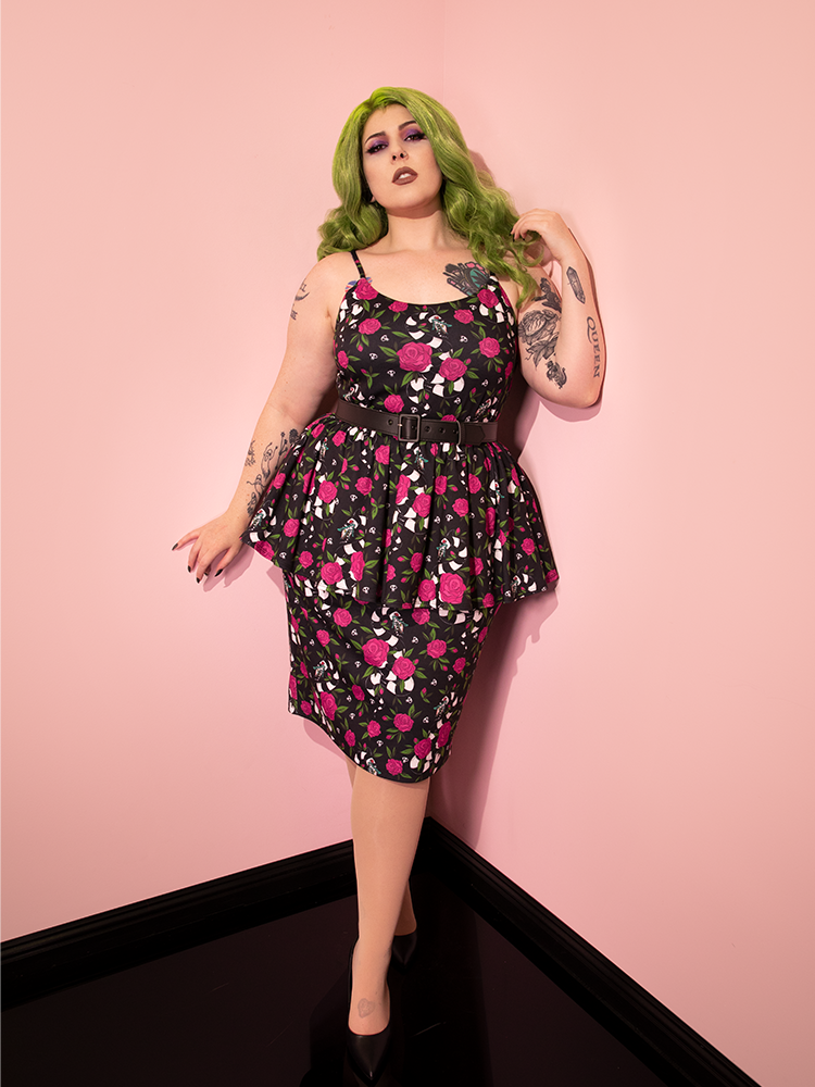 Green haired model wearing the BEETLEJUICE™ Sandworm & Roses Peplum Wiggle Dress in a pink room.