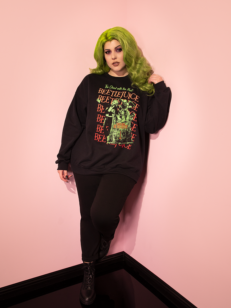 Full length photo of green-haired female model wearing the Tombstone Sweatshirt paired with black jeans and black ankle boots.