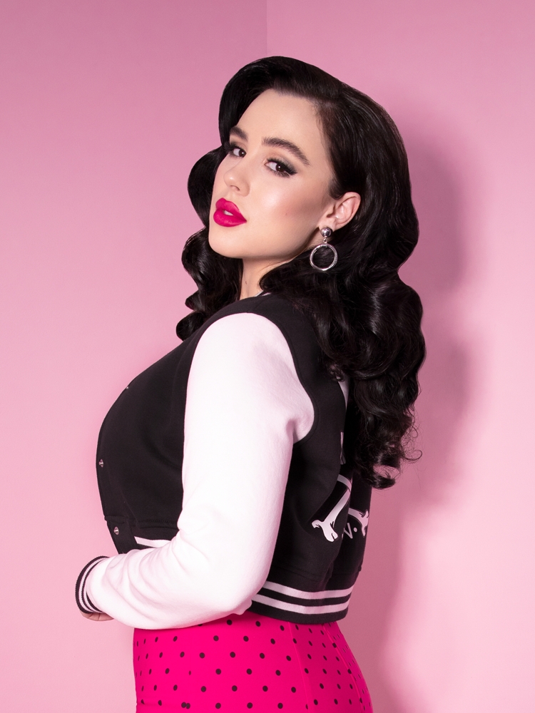 Rachel Sedory gazes longingly into the camera while sporting a Vixen Girl Gang Letterman Jacket and hot pink skirt with black polka dots.