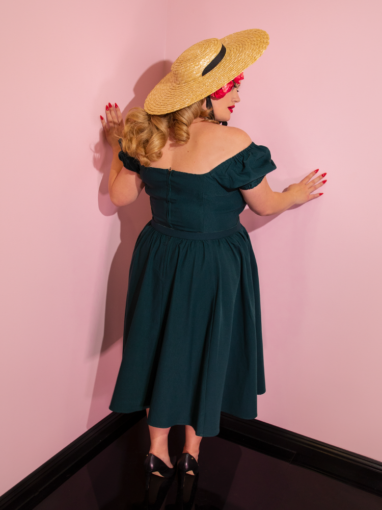 A back view of Blondie wearing a straw hat and flowers in her hair modeling the Vixen dress in hunter green.