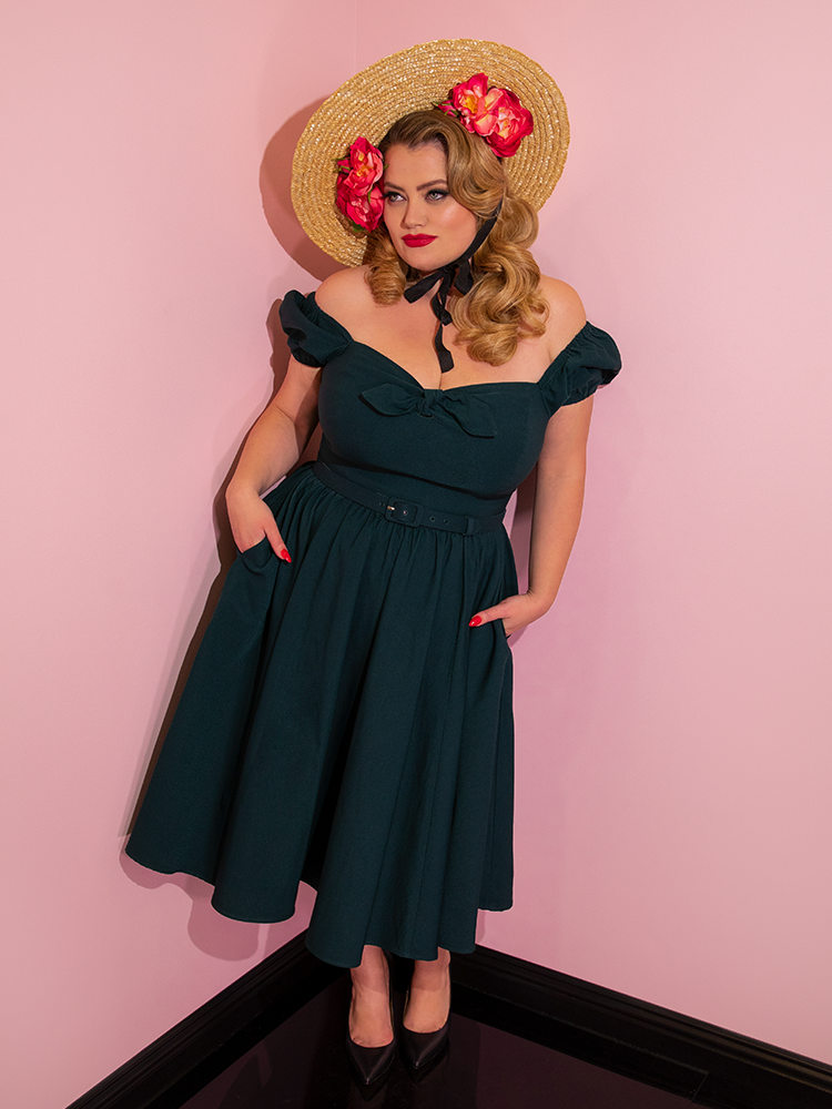 Blondie wearing a straw hat and flowers in her hair modeling the Vixen dress in hunter green with her hands in her pockets.