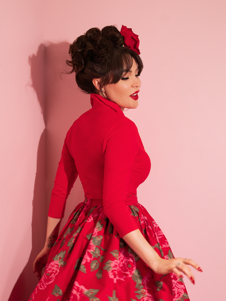 Micheline Pitt turned to the side provides a side shot of the Vixen Top in Ravishing Red with pink rose skirt.