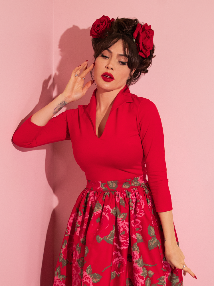 Micheline Pitt posing in the Vixen Top in Ravishing Red tucked into a pink rose skirt with rose hair clips on either side of her head.