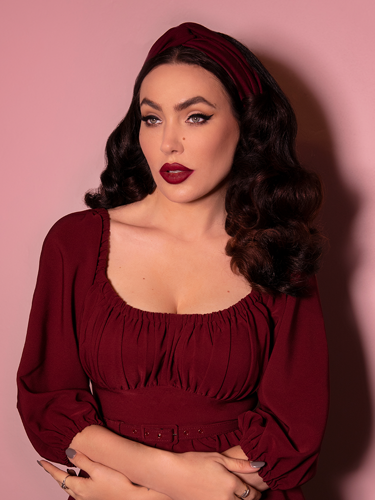 A closeup shot of Micheline Pitt looking past the camera while modeling the knot headband in burgundy paired with a matching dress.