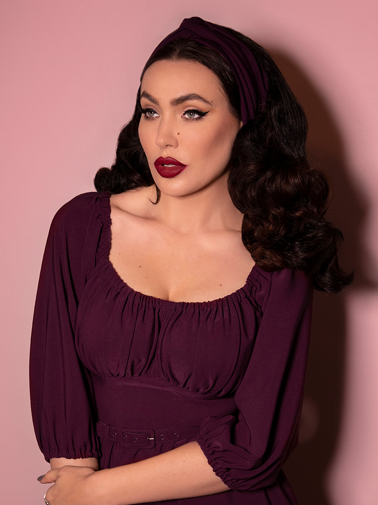 A closeup shot of Micheline Pitt looking past the camera modeling the vintage style knot headband in eggplant paired with a matching dress.