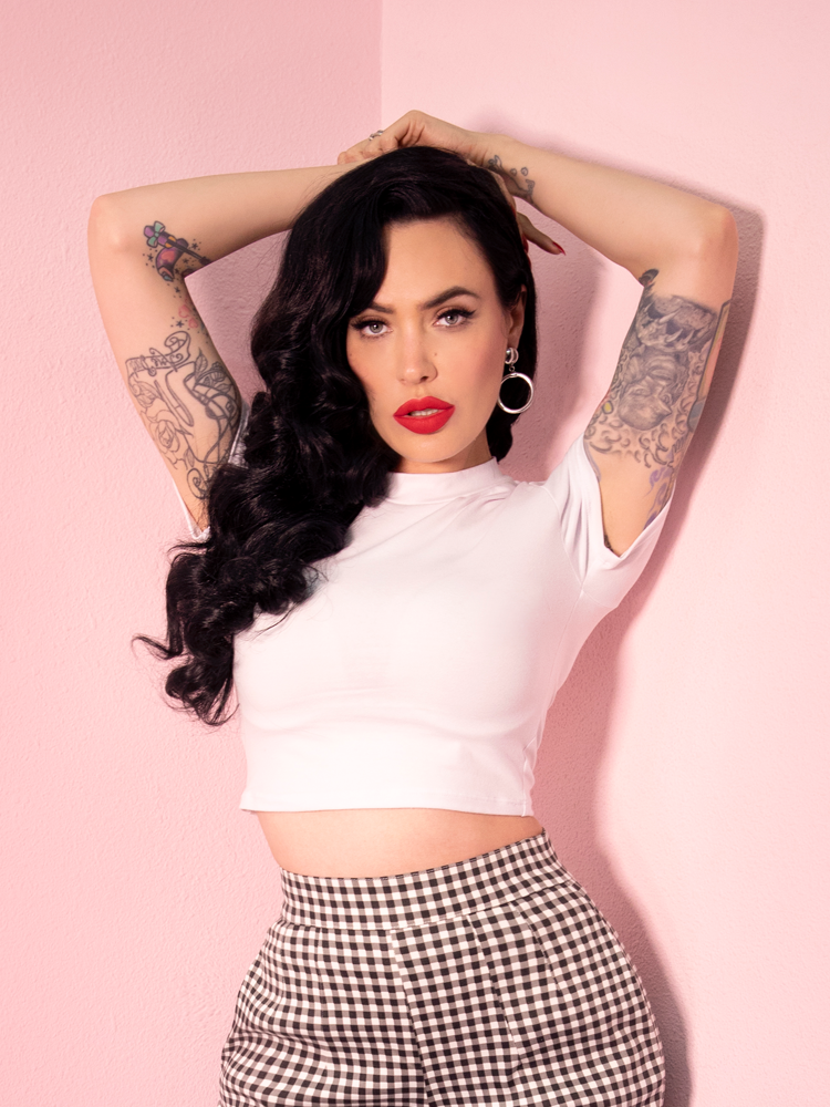 A closeup of Micheline Pitt with her hands behind her head modeling the Bad Girl crop top in white paired with gingham cigarette pants.