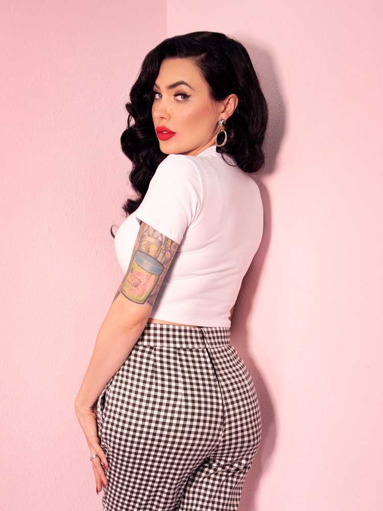 A back view of Micheline Pitt looking over her shoulder modeling the Bad Girl crop top in white paired with gingham cigarette pants.