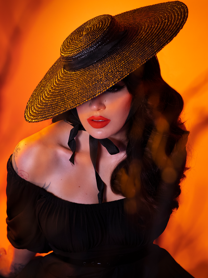 Micheline Pitt, standing against a blood orange background, wearing the Regular Vintage Sun Hat in Black from Vixen Clothing.