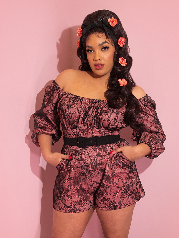 A closeup of Ashleeta with pink flowers in her hair and her hands in her pockets modeling the Vacation Playsuit in peach and black lace.