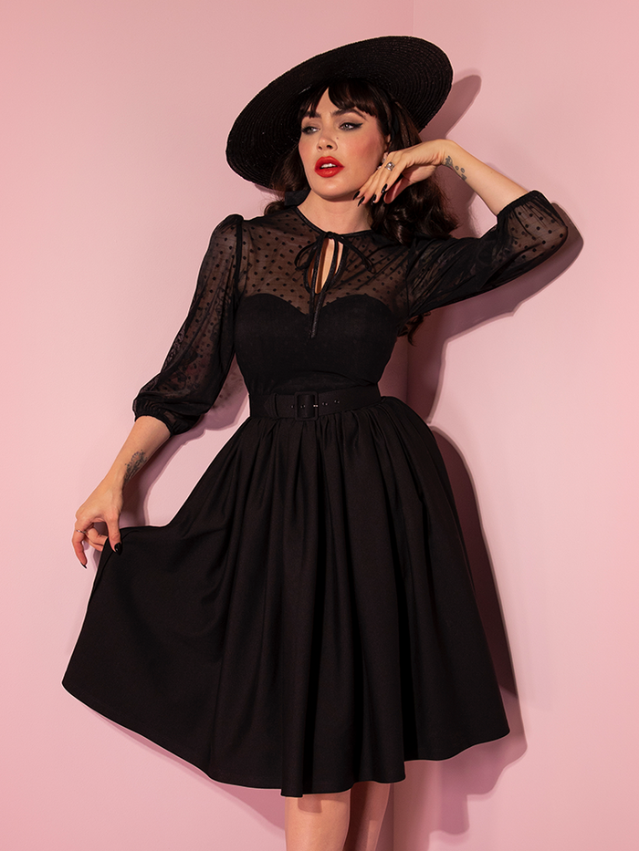 Micheline Pitt posing with one hand on her chin and the other gently pulling out the skirt section of the Frenchie Swing Dress in Black.