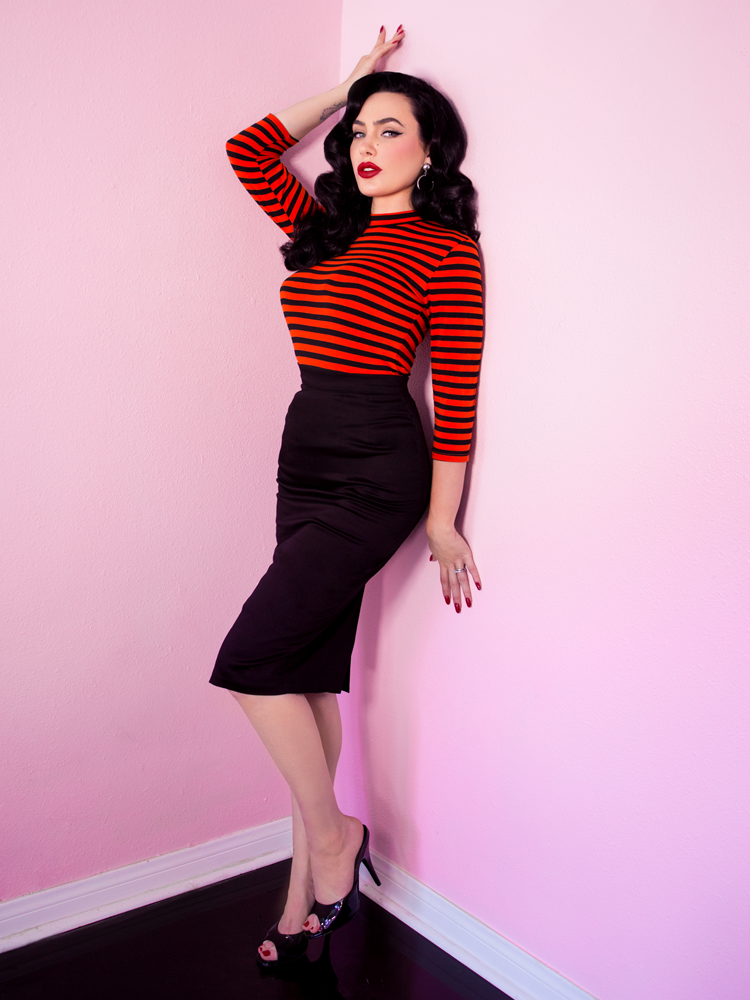 Full length shot of Micheline Pitt in a retro style pencil skirt, orange and black long sleeve top and black heeled shoes.