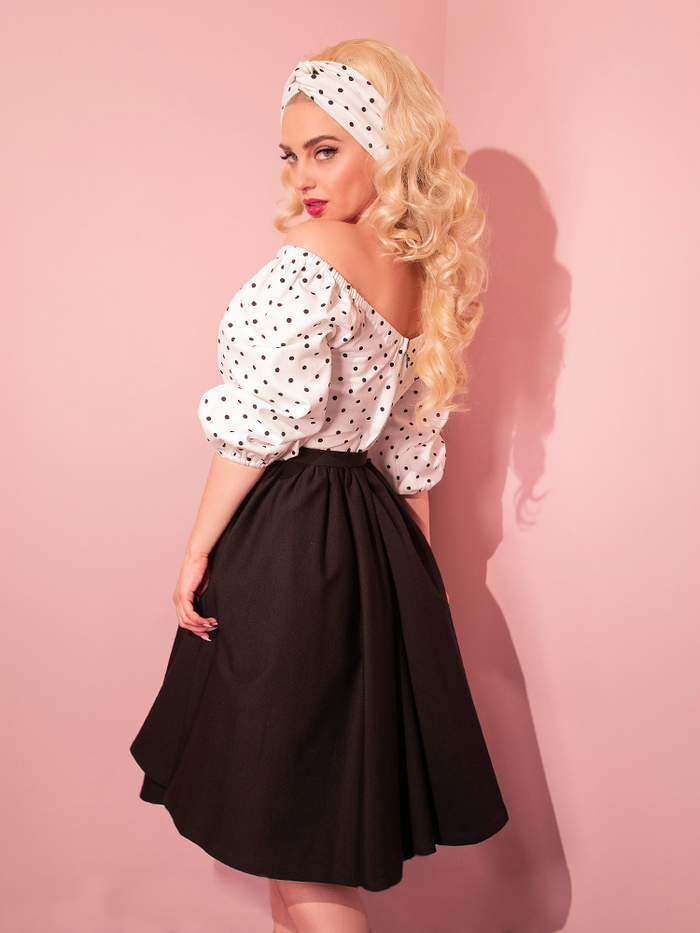 Platinum blonde model wearing the Daydream Swing Dress in Black Polka Dot turns to the side.