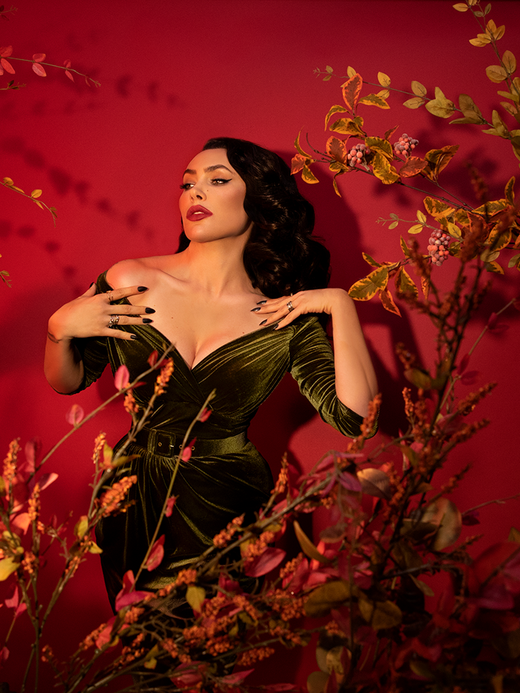 Stylized shot of Micheline Pitt in the Starlet Wiggle Dress in Olive Green posing in front of a blood red background and amongst yellow and green leaved branches.