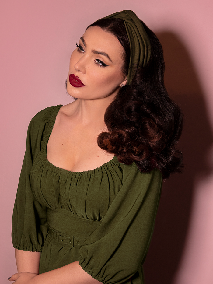 Looking off camera, Micheline Pitt models the vintage style knot headband in olive green paired with a matching dress.