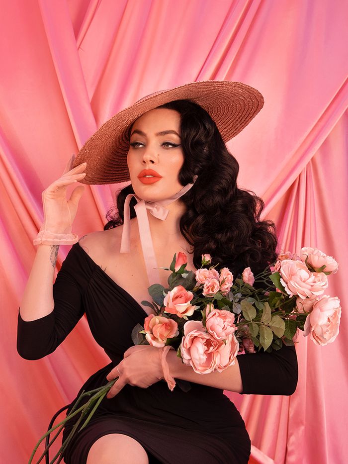 Micheline Pitt sitting in front of a pink backdrop, holding pink roses while wearing a vintage style black dress and the Vintage Sun Hat in Blush Pink.