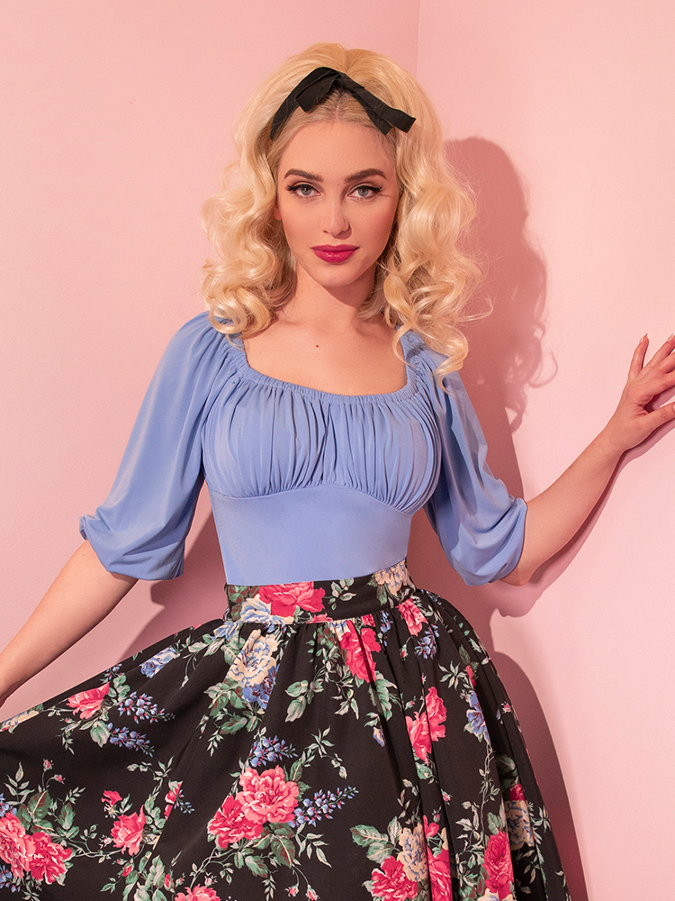 Platinum blonde model wearing the Vacation Blouse in Sunset Blue tucked into a black, floral print skirt. 